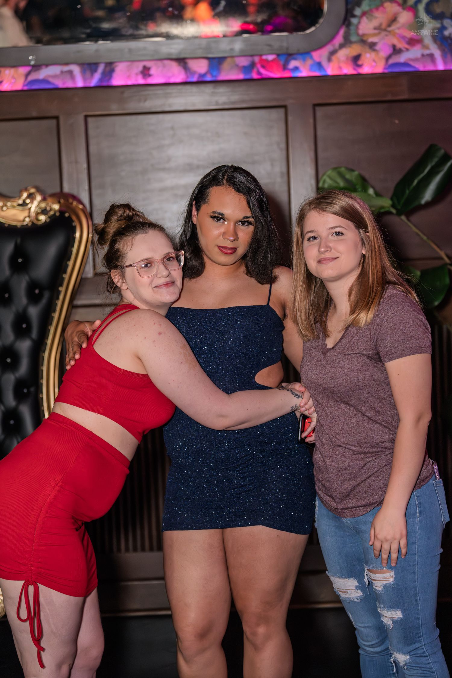 Three women pose together at a nightclub, one in red, one in a navy sparkle dress, one in a mauve top.