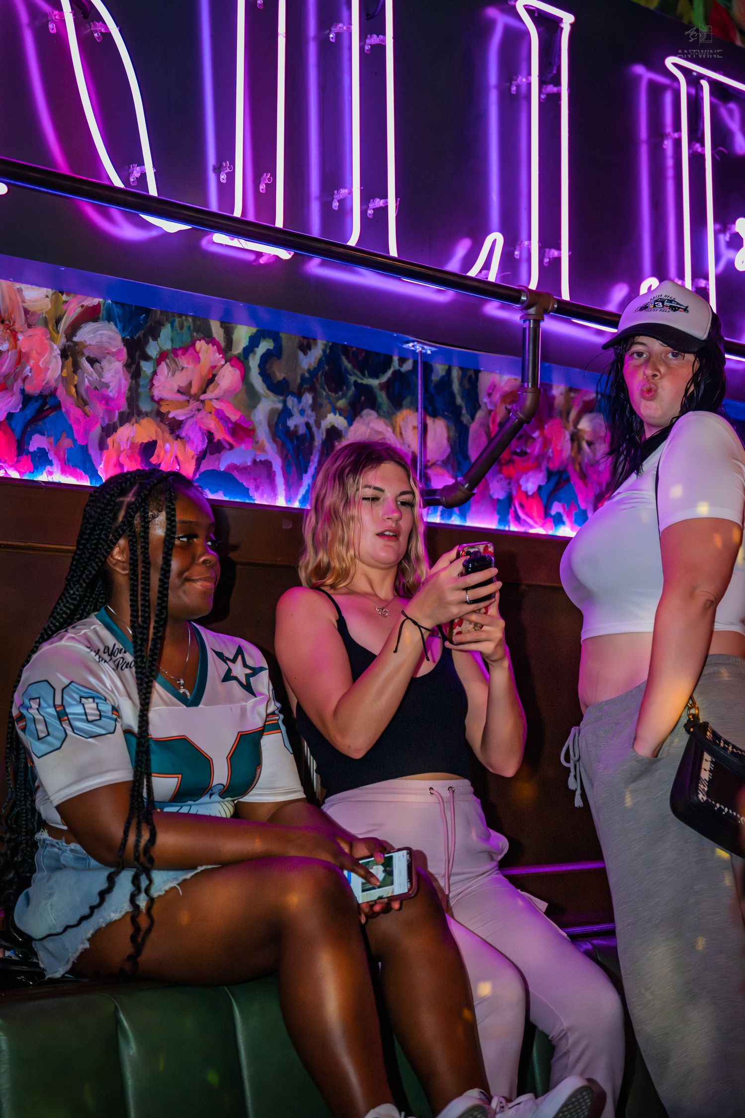 Three women socializing in a nightclub booth beneath glowing purple neon signs and floral artwork.