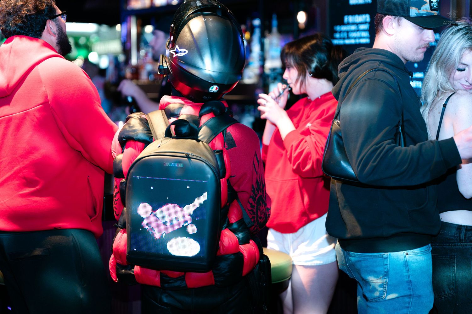 People in red hoodies with VR gaming backpacks mingling at a busy nightclub event.