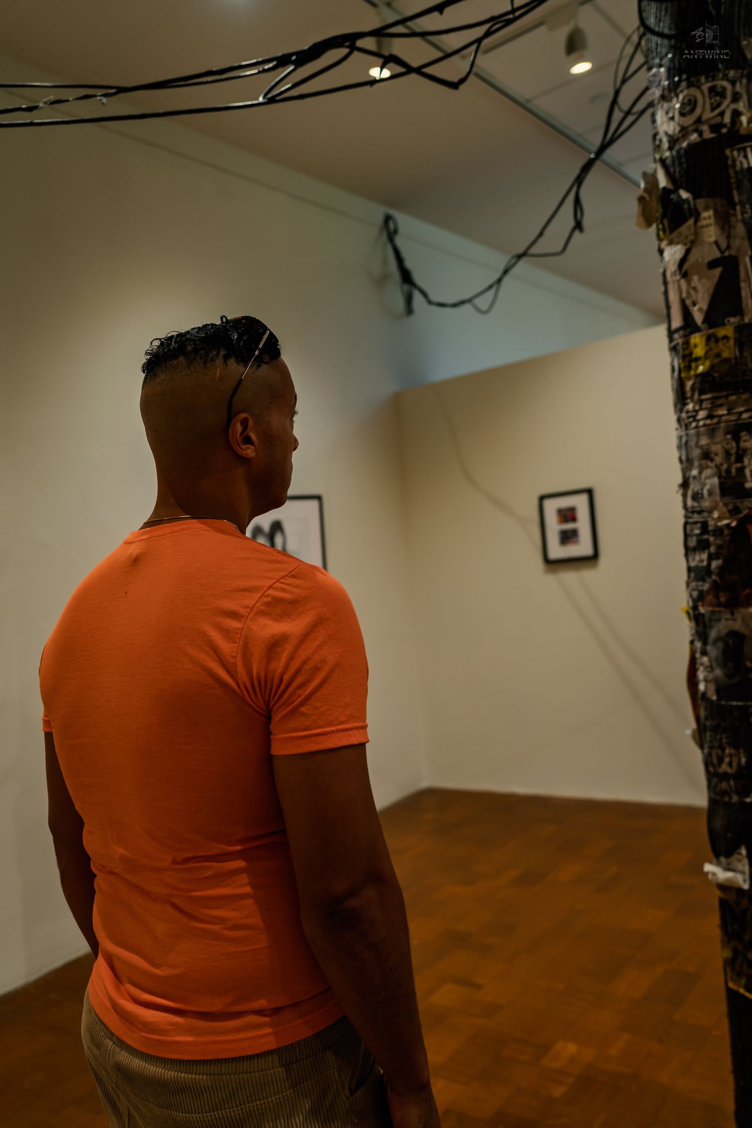 A man in an orange t-shirt views contemporary art installation with hanging wires in a gallery.