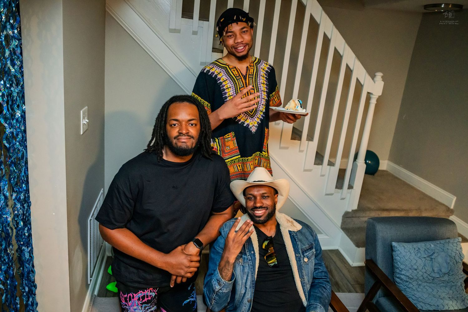 Three young men smile together near a staircase, one wearing a colorful dashiki shirt at a party.