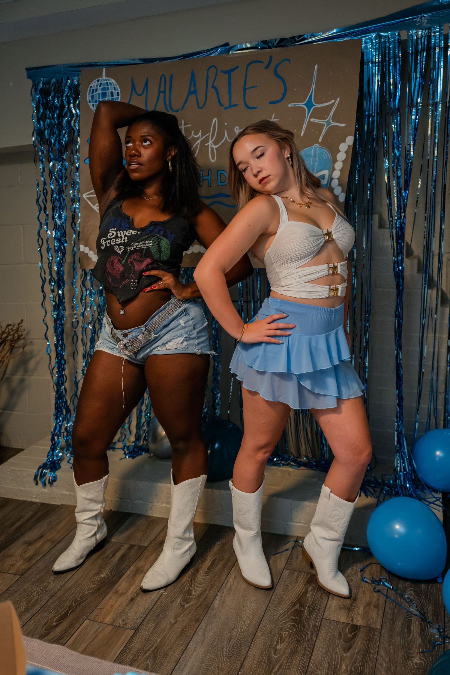 Two women in white cowboy boots pose together at a blue-themed 21st birthday party backdrop.