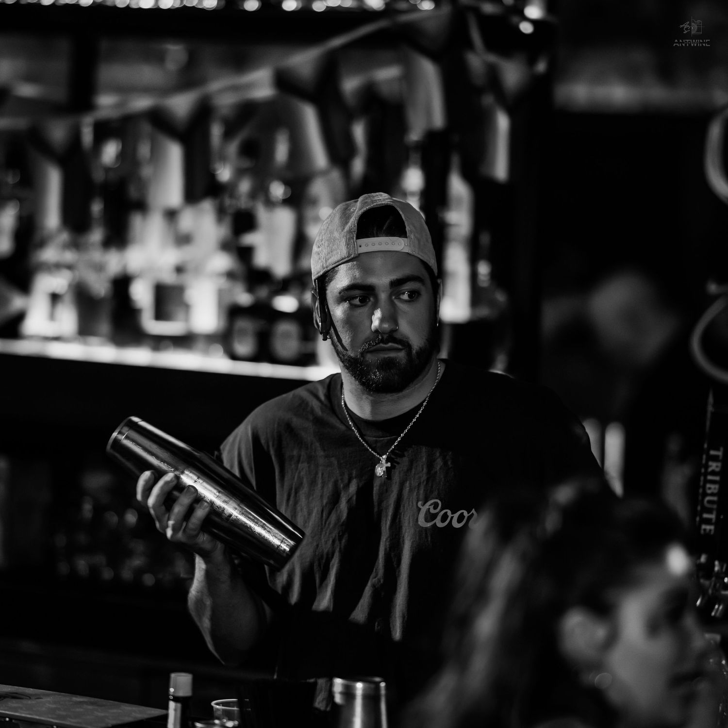 Black and white photo of male bartender shaking cocktail at a well-stocked bar.