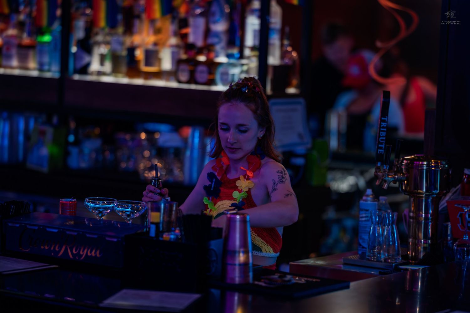Female bartender with tattoos preparing drinks at a dimly lit, colorfully stocked bar.