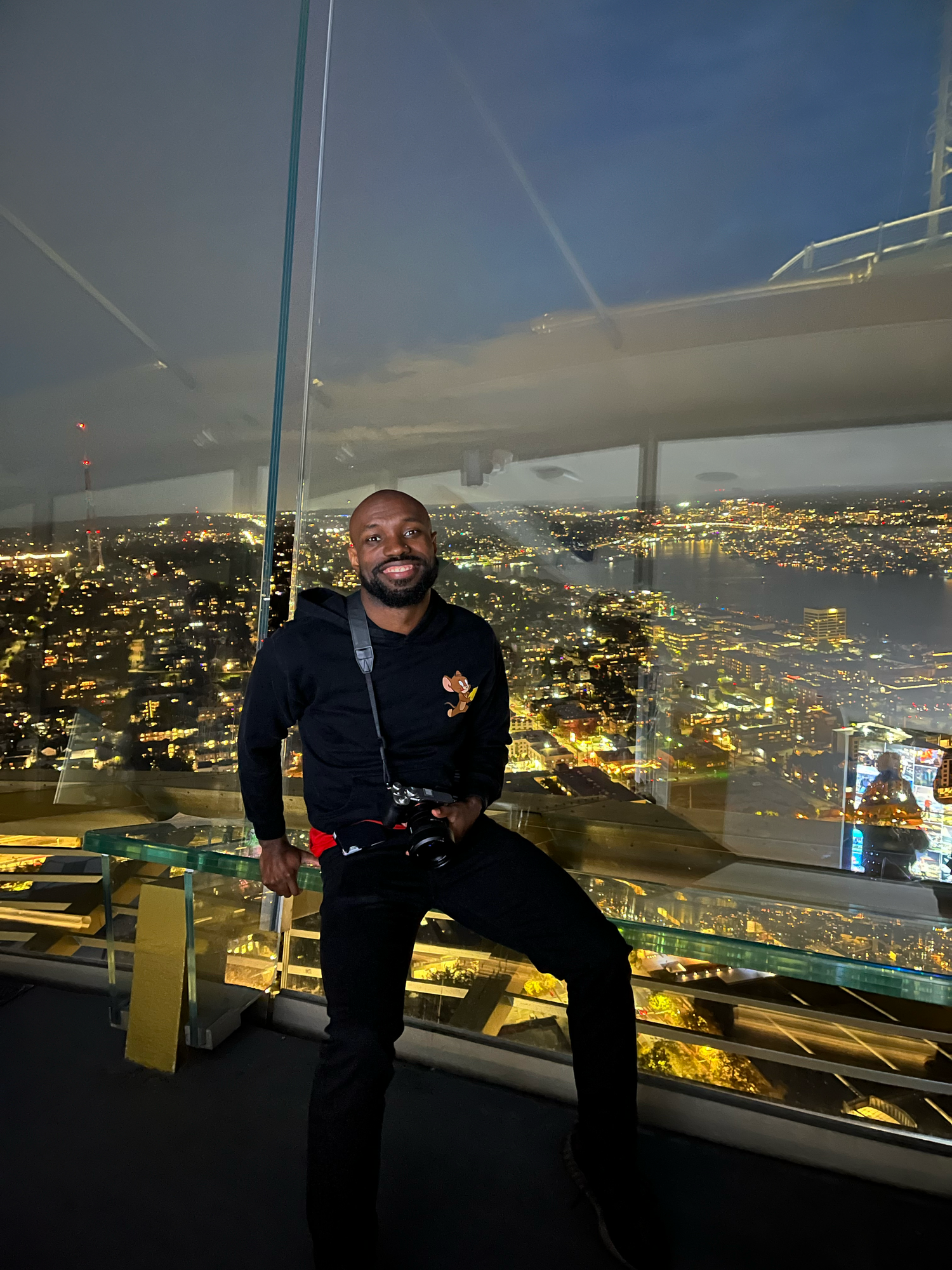 Man smiling on glass observation deck at night with illuminated city skyline and waterfront visible below.
