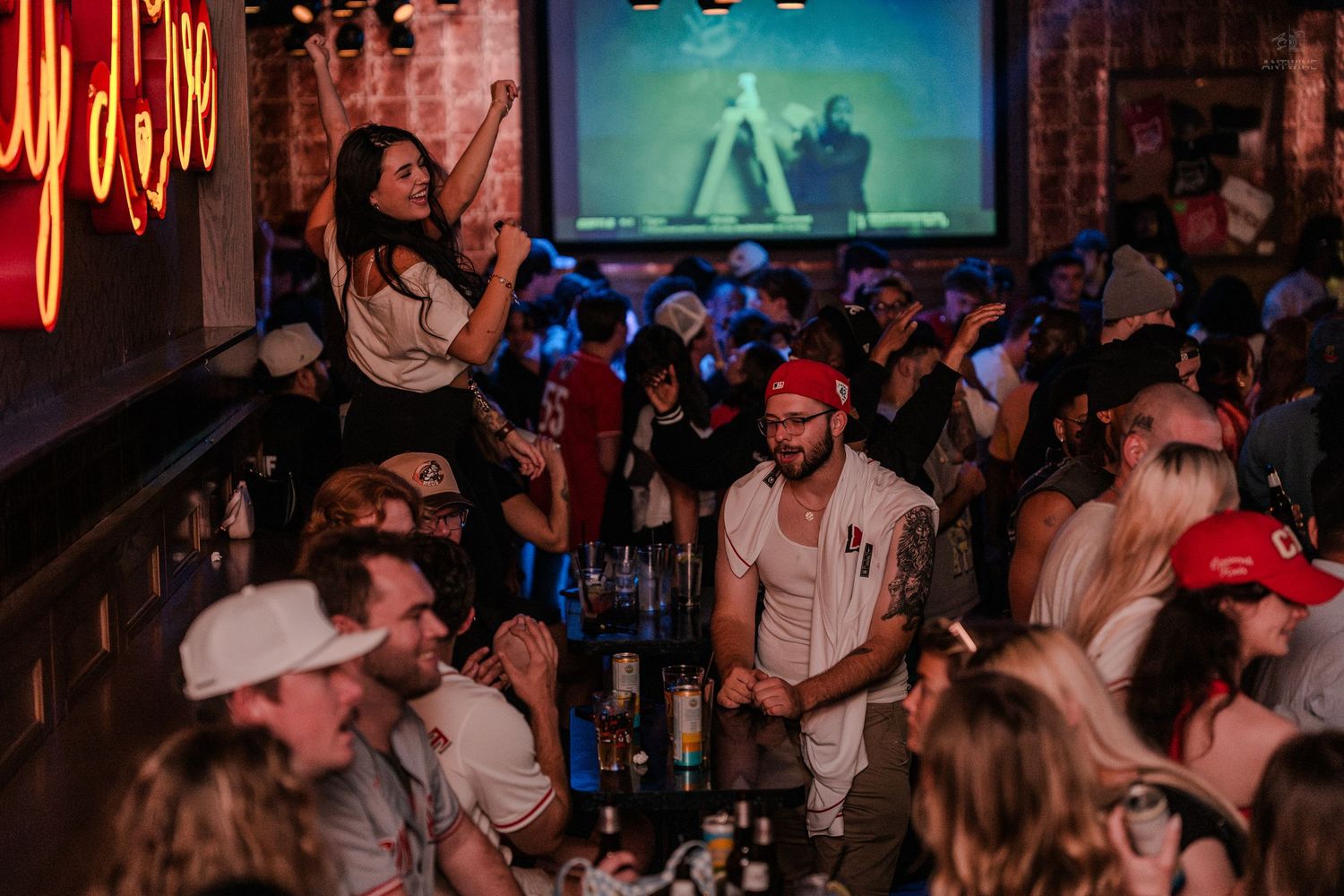 Energetic crowd enjoys a lively bar event with neon signs and a large screen displaying video.