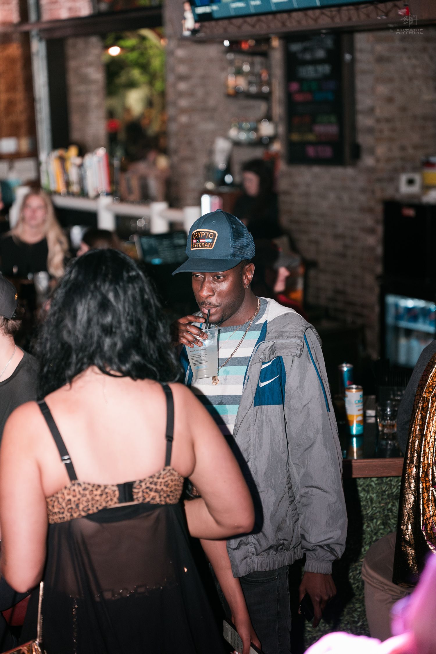 Man in grey Nike jacket and blue cap sipping a drink at a lively bar or nightclub event.