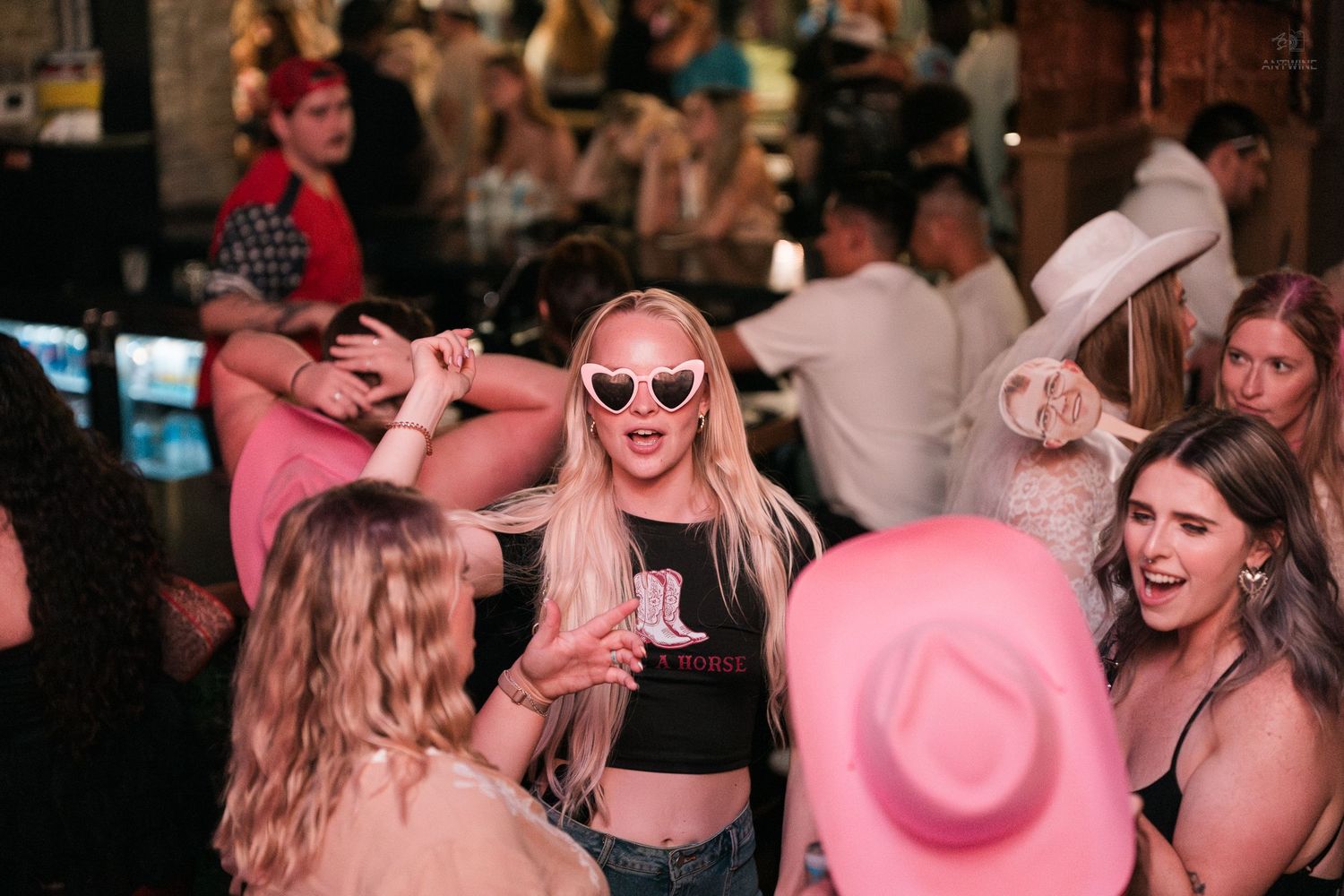 Blonde woman in heart sunglasses dances with friends wearing pink cowboy hats at a lively party.