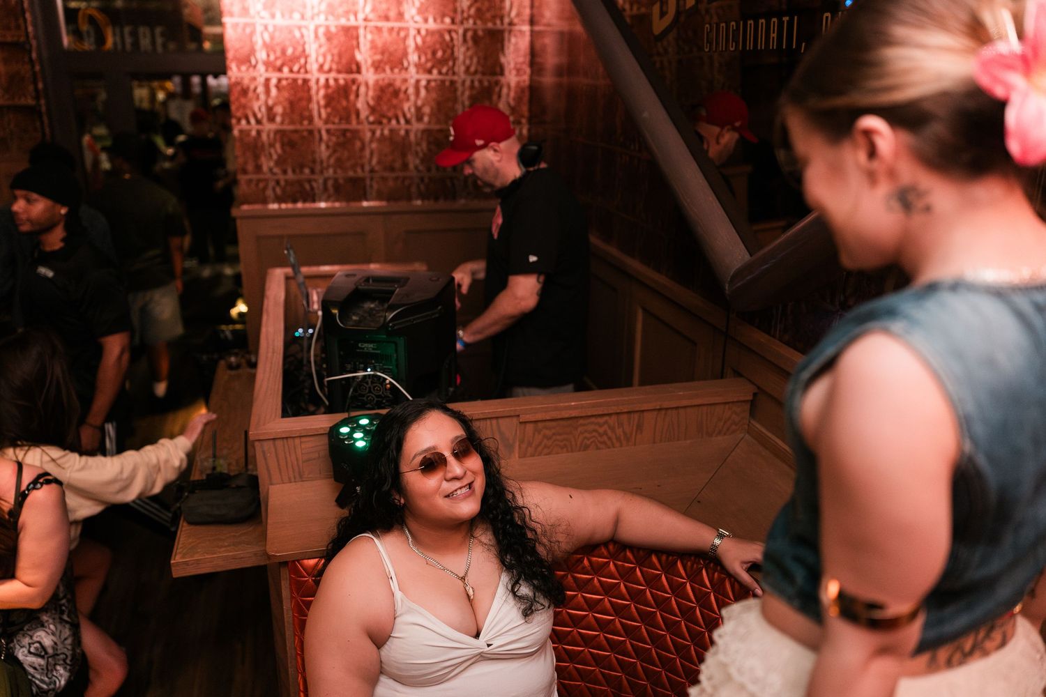 A woman wearing headphones smiles up at a friend near a DJ booth inside a dimly lit bar.