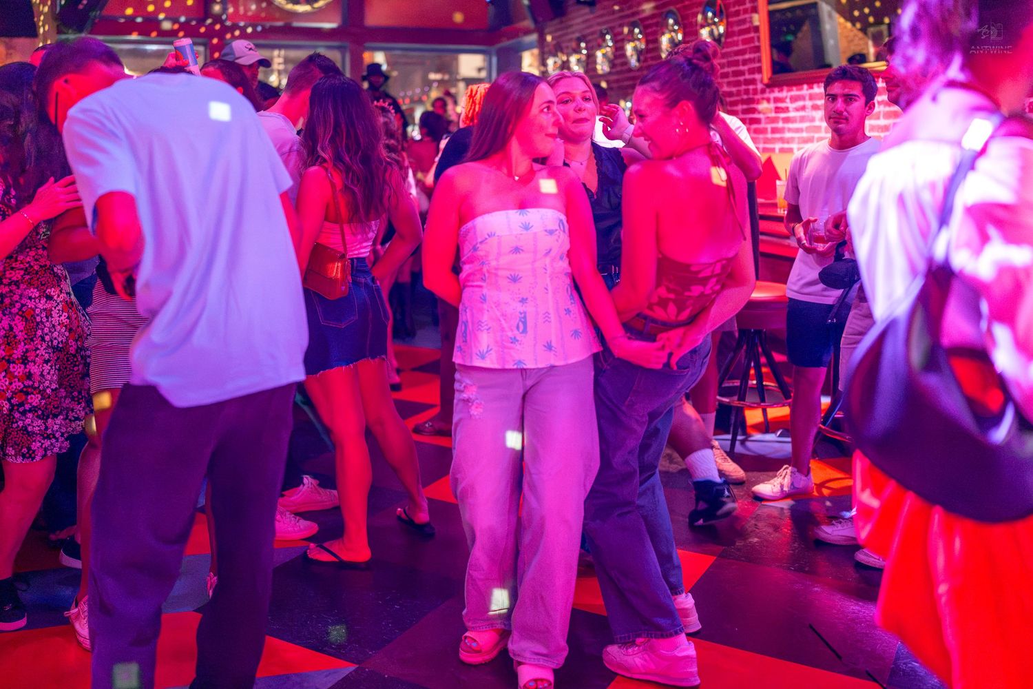 Young adults dancing together under pink and red lights in a lively nightclub or bar setting.