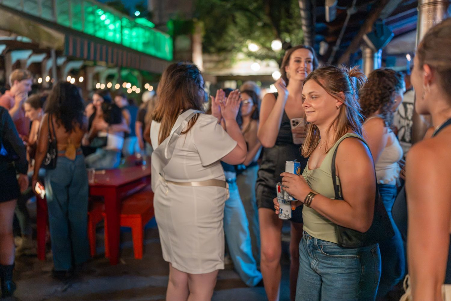 Women socializing and high-fiving at an outdoor bar or nightlife event with string lights in the background.