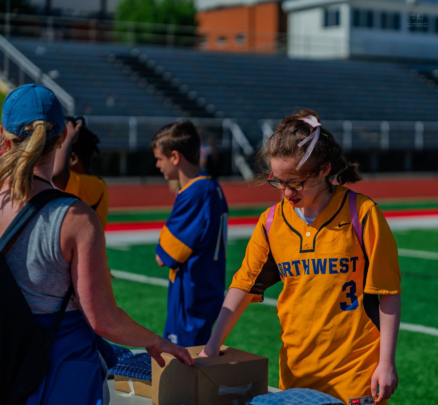 Northwest softball player #3 receives items at a stadium table during a team event on a sunny day.