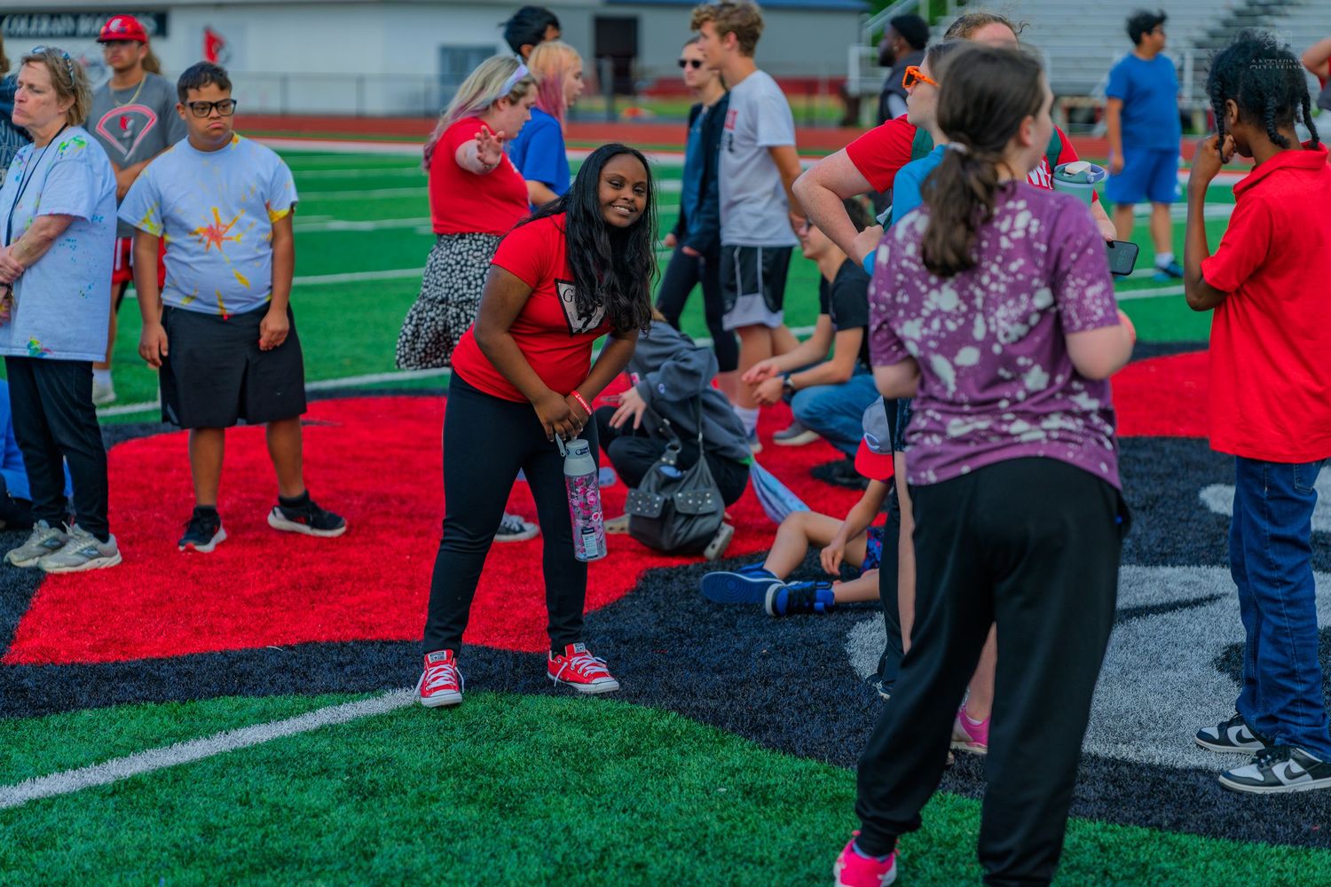 Students socializing on a red turf athletic field during an outdoor school event on a sunny day.