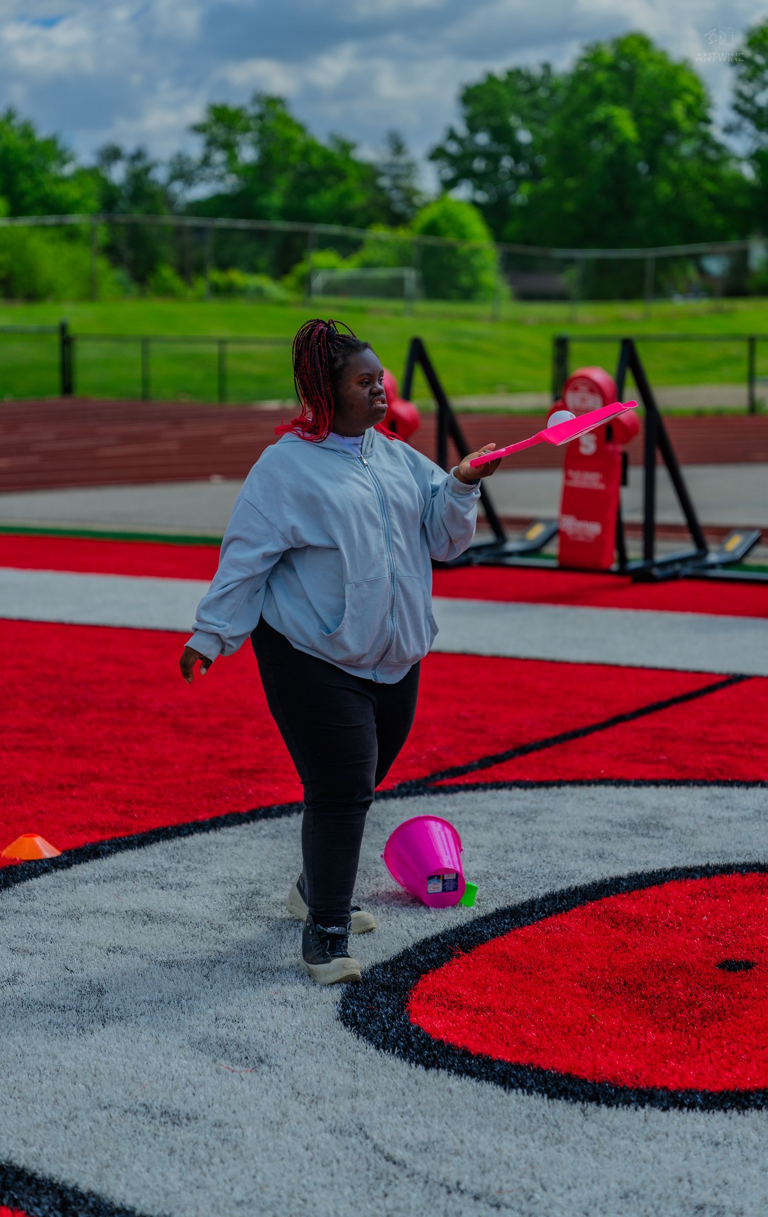Woman in gray hoodie and black leggings holds pink toy on red track field outdoors on cloudy day.