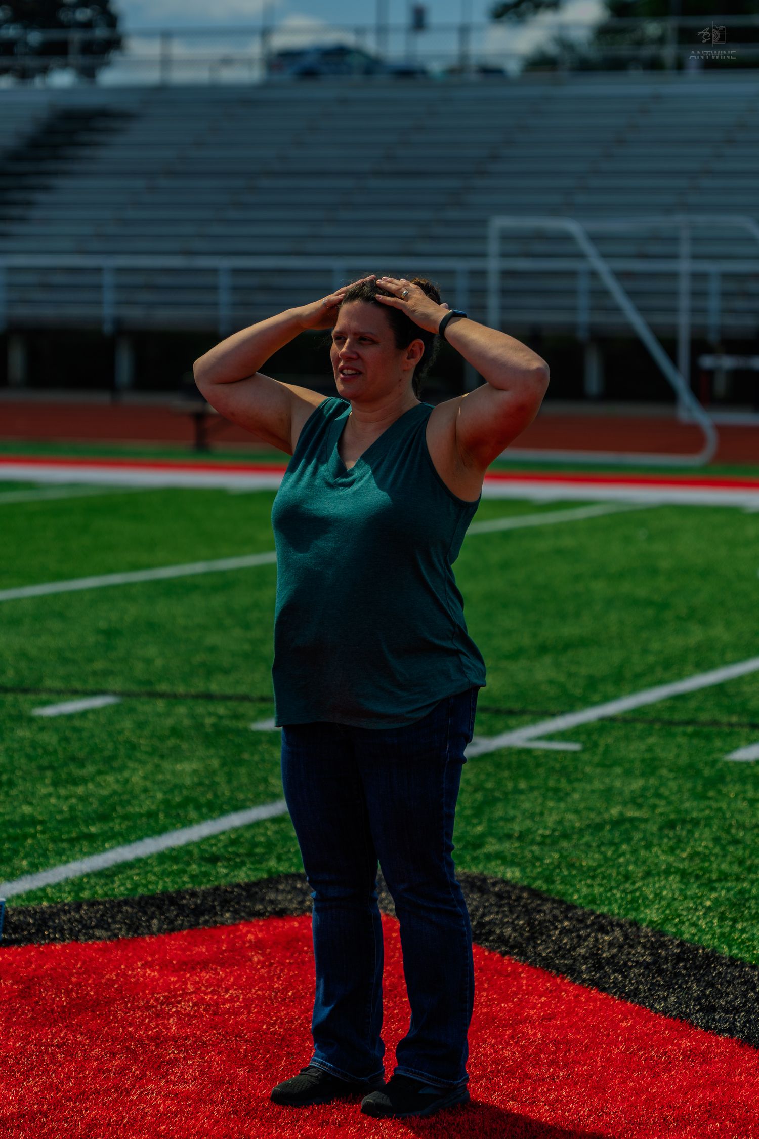 Woman in teal tank top stands on red zone of football field with hands behind head, stadium bleachers in background.