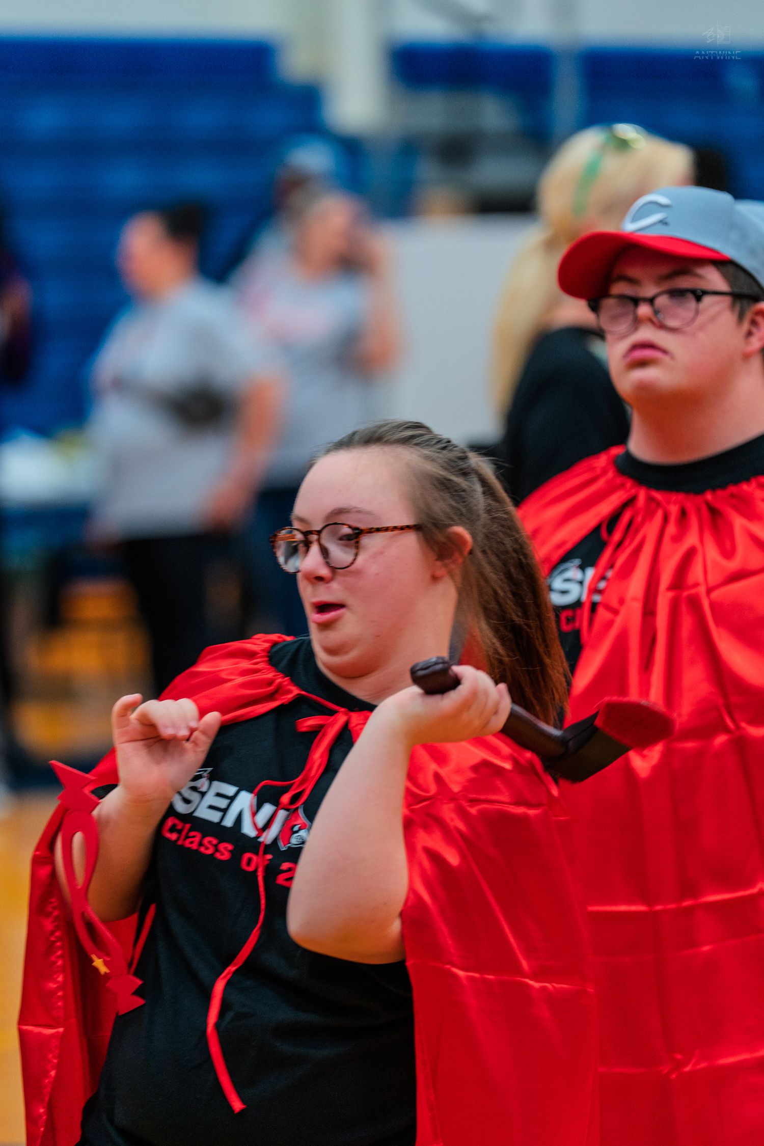 Two high school seniors in red capes and black Senior Class of 2023 shirts celebrate at a gymnasium event.