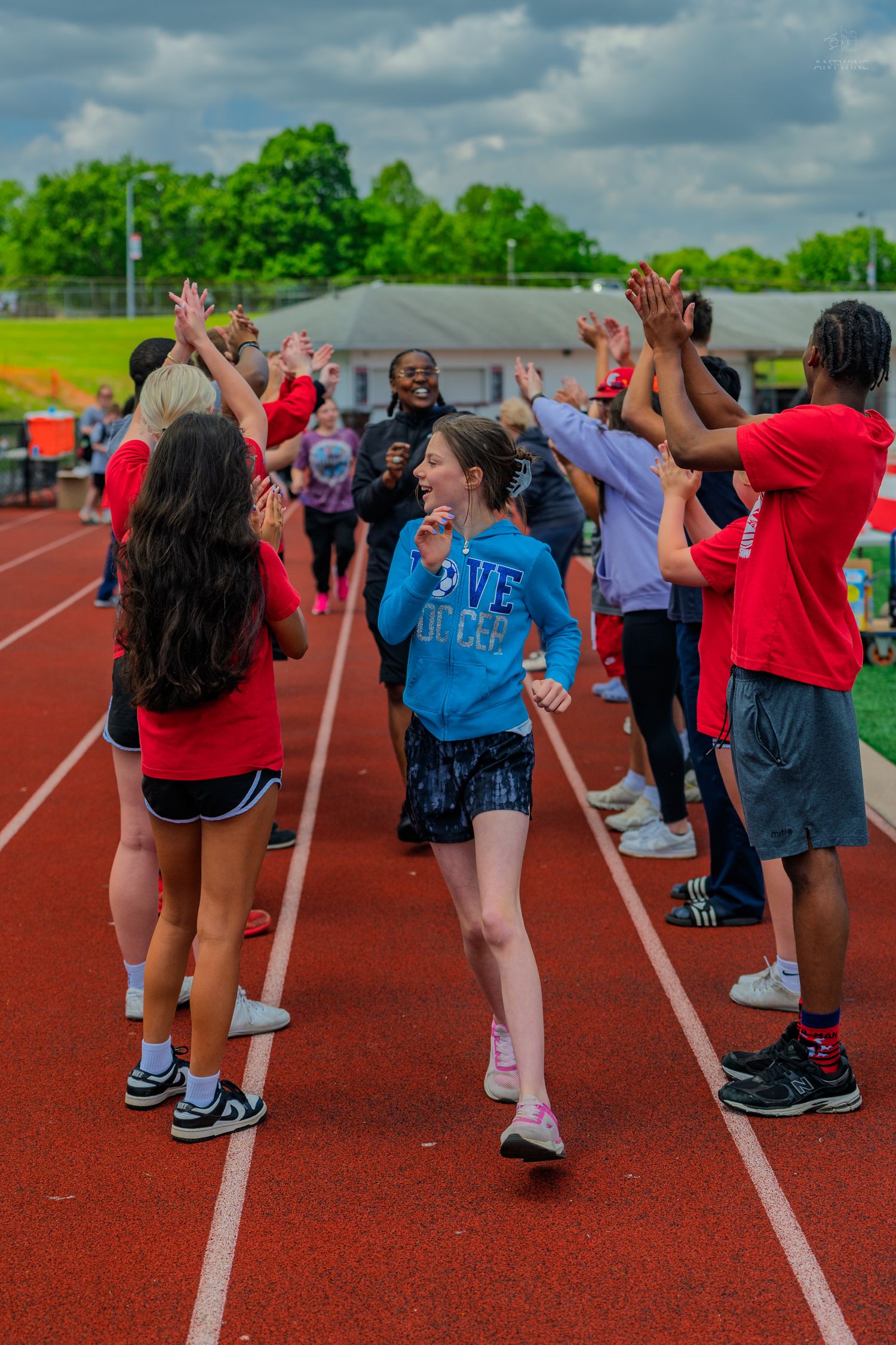 Students running through a high-five tunnel on a red track during an outdoor school sports event on a cloudy day.