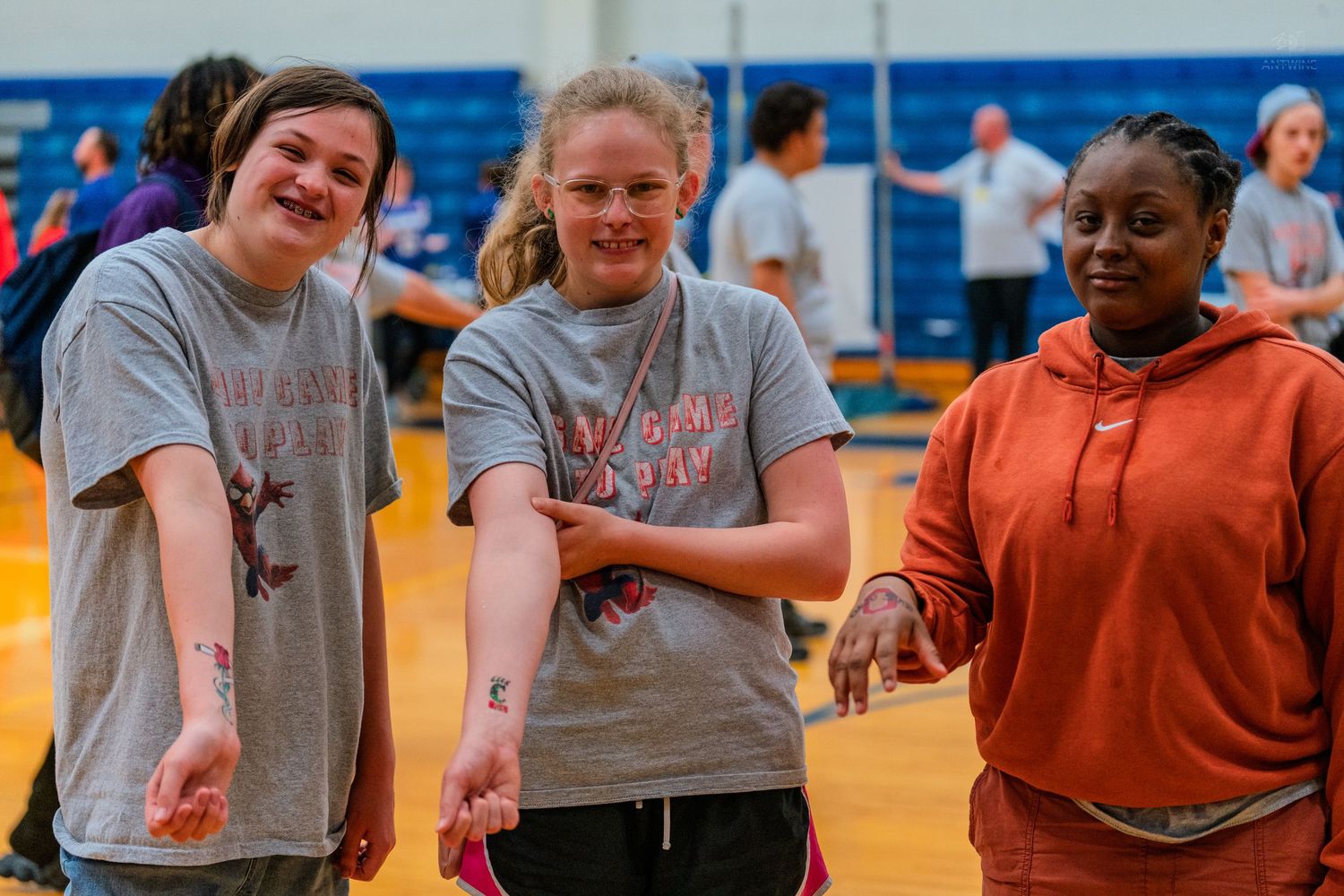 Three smiling girls holding hands on a gymnasium floor during a school sports event.