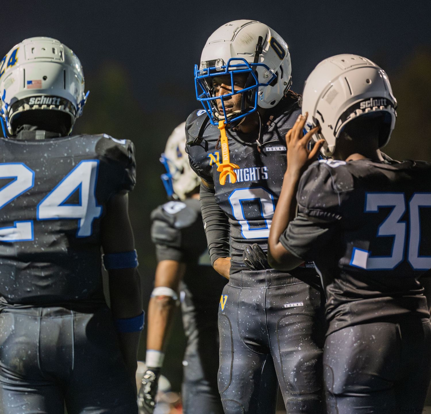 Three football players in dark Knights uniforms huddle together during a night game.