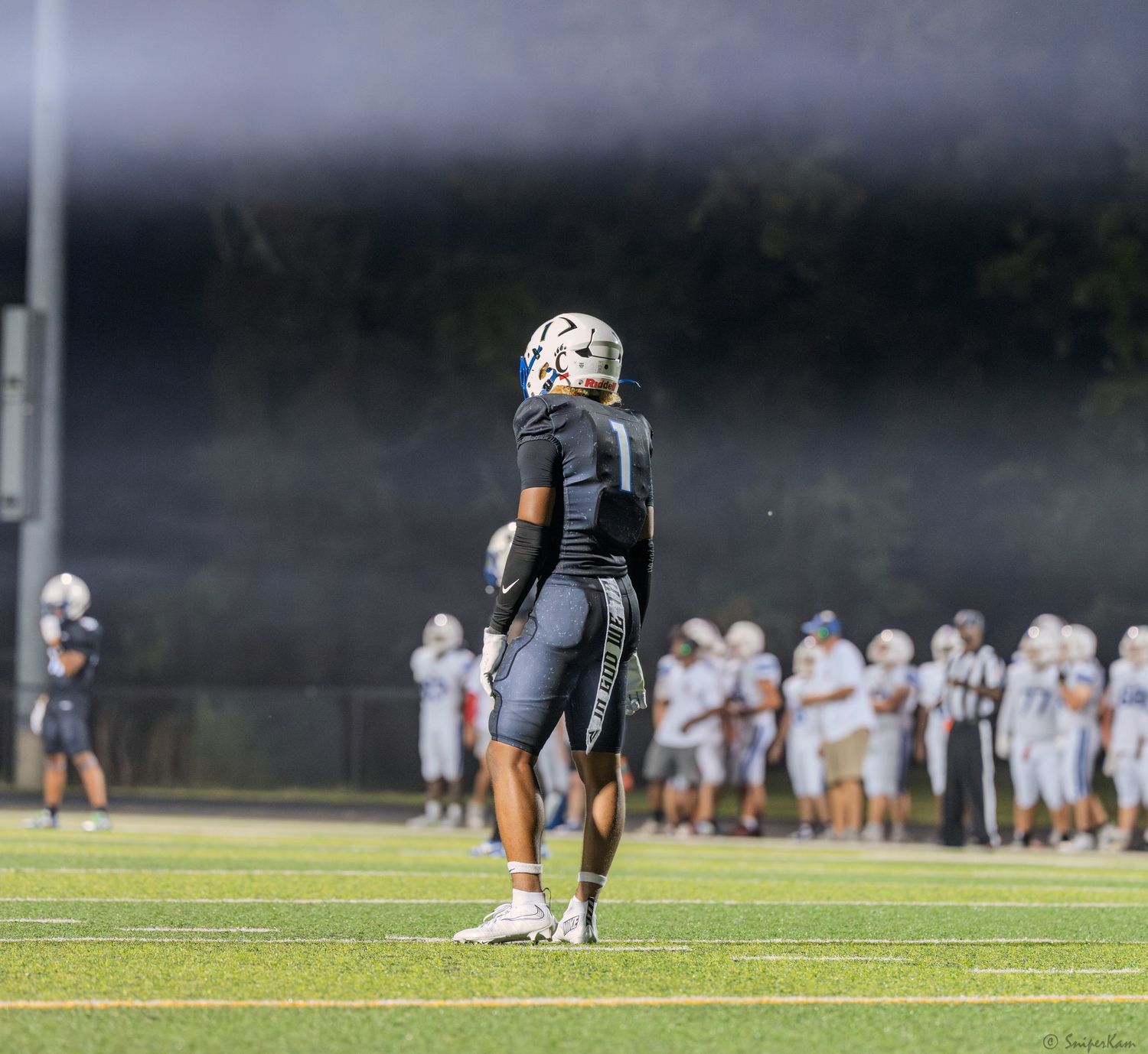 High school football player in black uniform stands alone on field under bright stadium lights during a game.