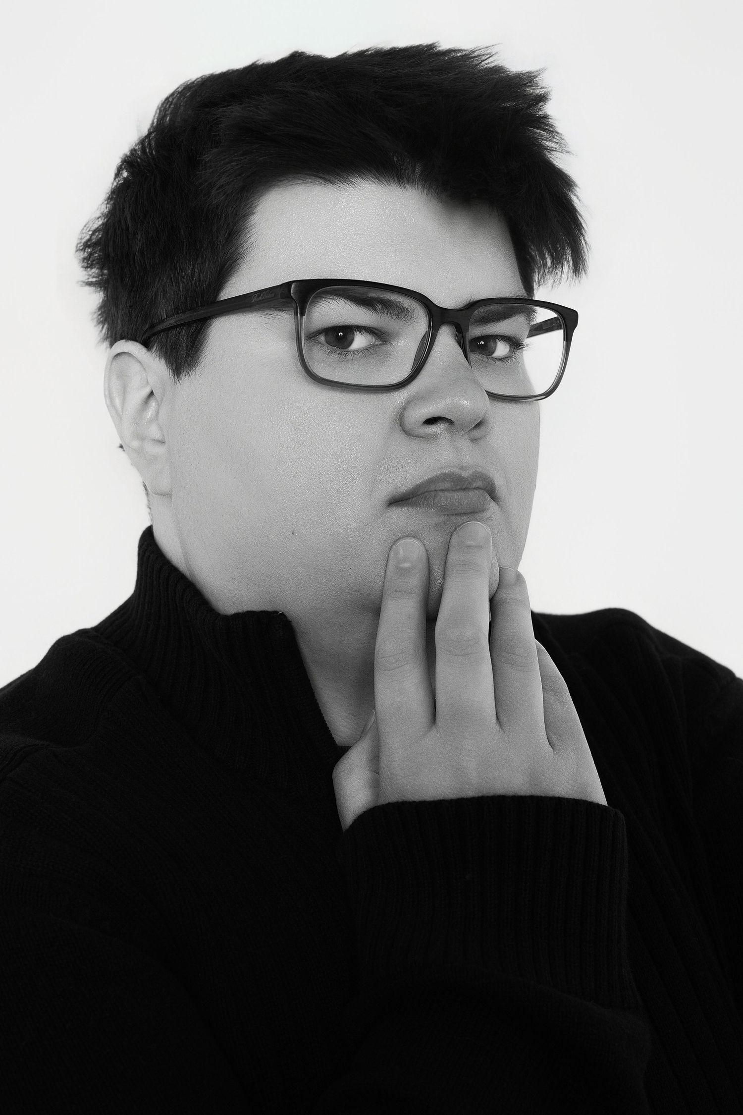 Black and white portrait of a young person wearing glasses with hand raised thoughtfully to chin.