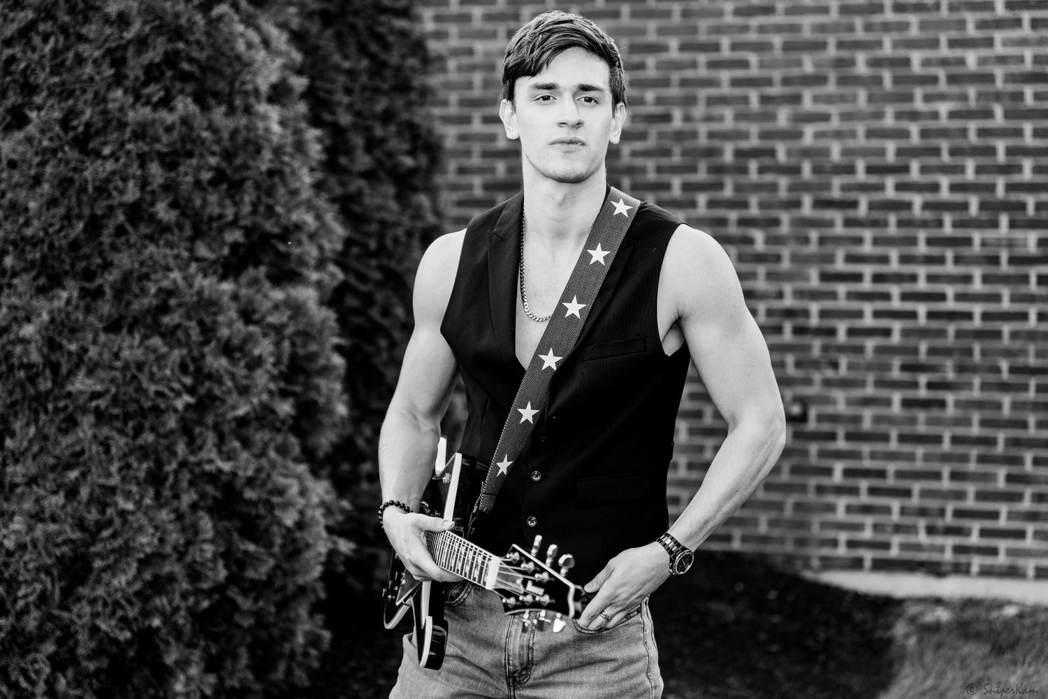 Young male guitarist in sleeveless vest holding guitar outdoors near brick wall in black and white.