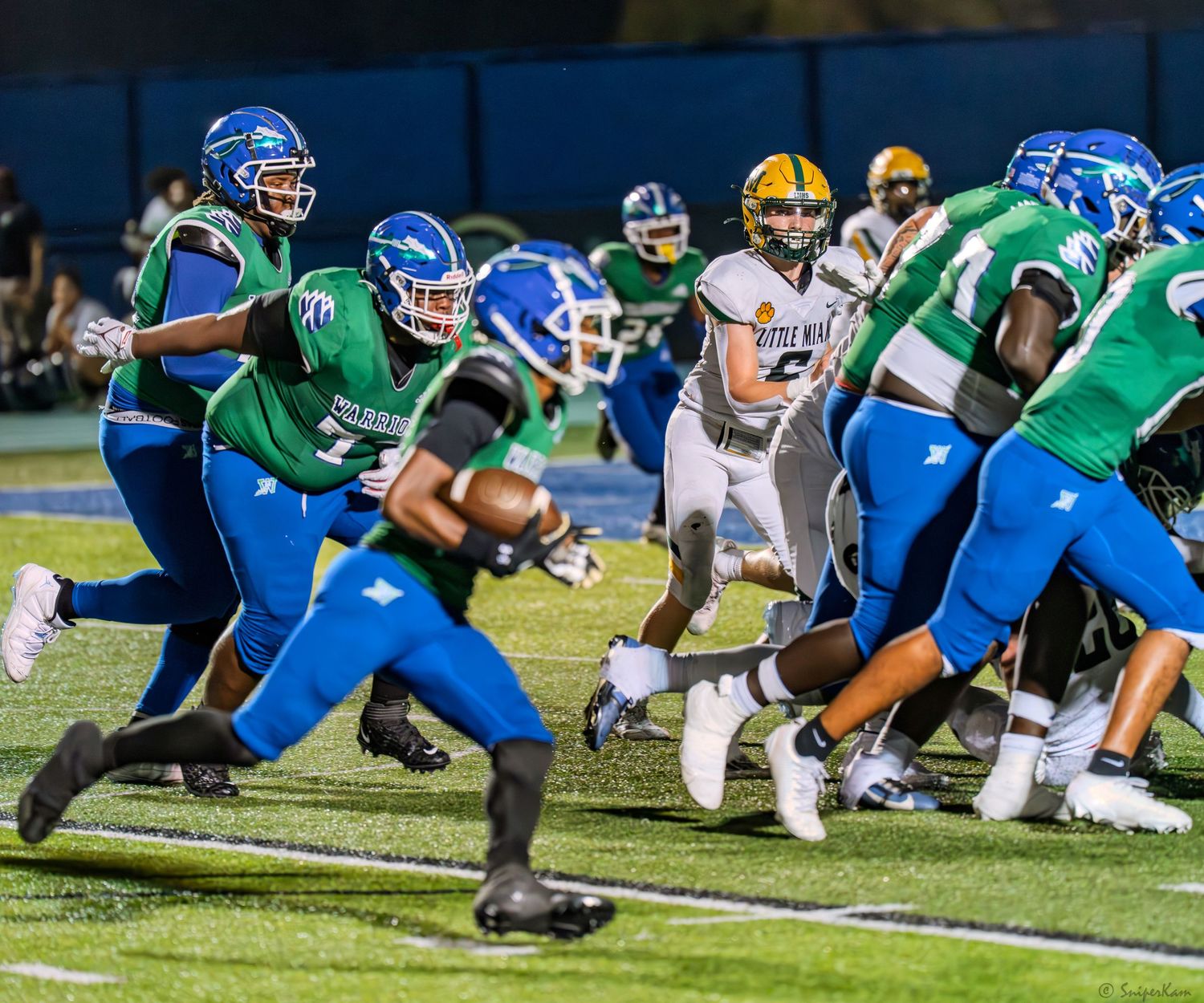 High school football players in blue and green uniforms execute a running play during a night game.