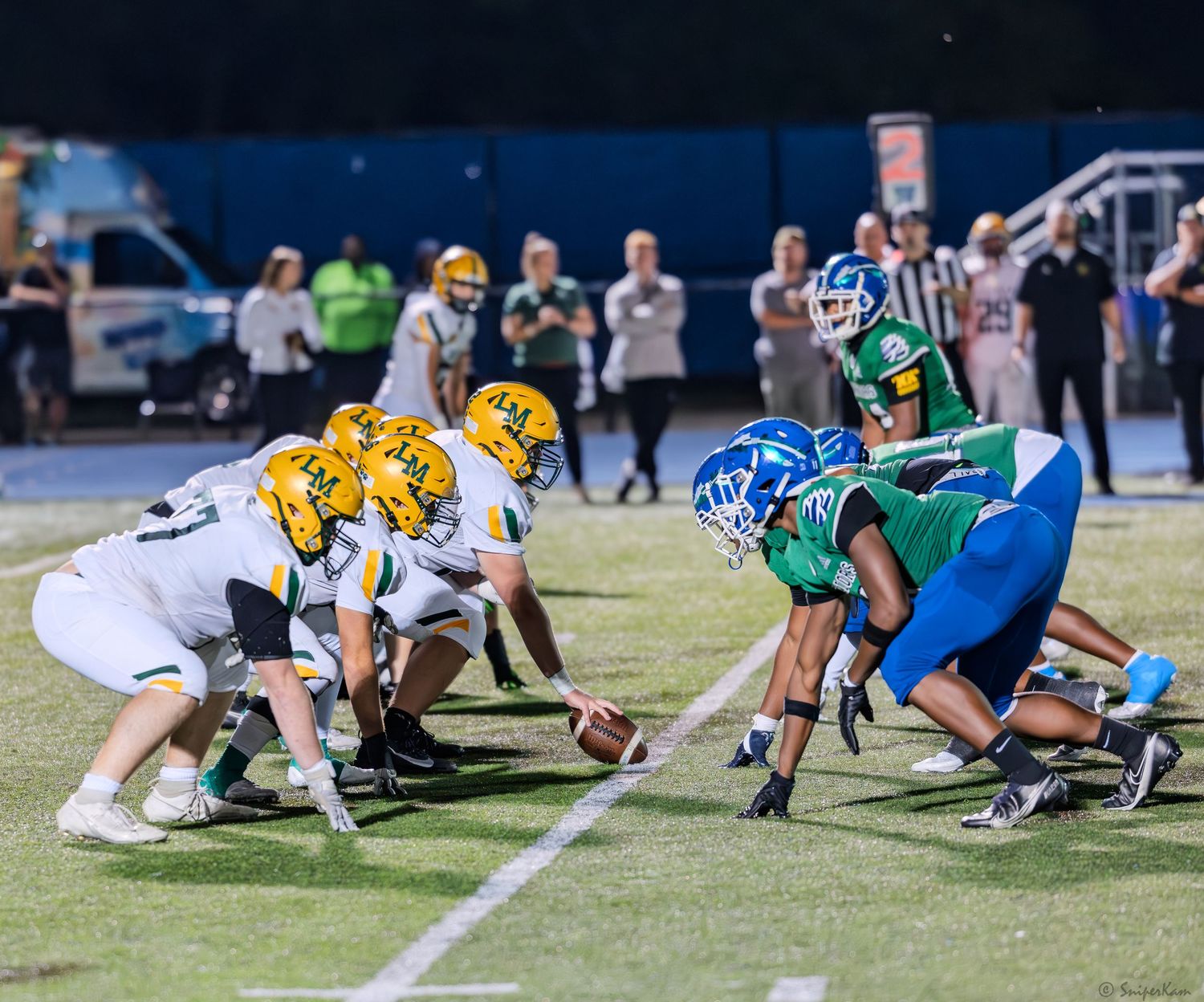 High school football teams line up at the line of scrimmage during a night game under stadium lights.