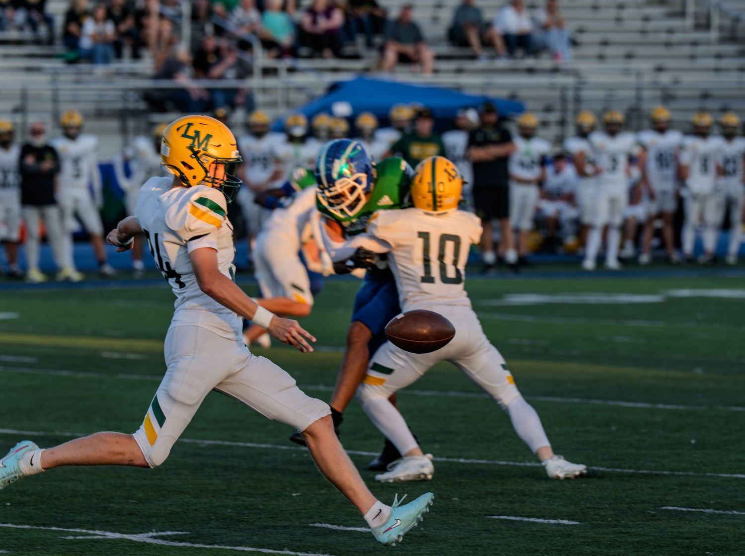 High school football players in gold uniforms execute a handoff play during a daytime football game.