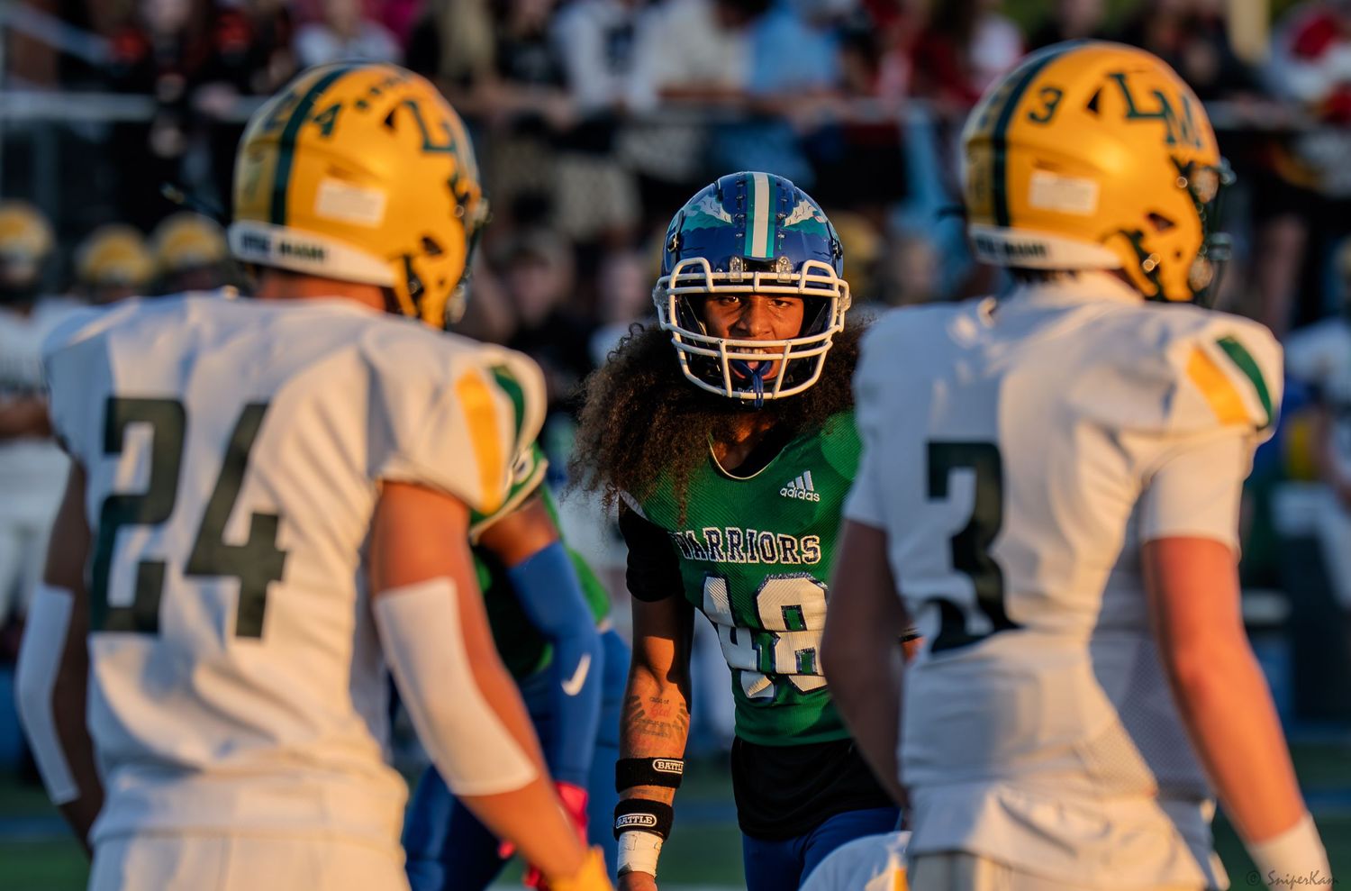 Green Warriors football player #48 faces off against yellow-helmeted opponents #24 and #3 on the field.