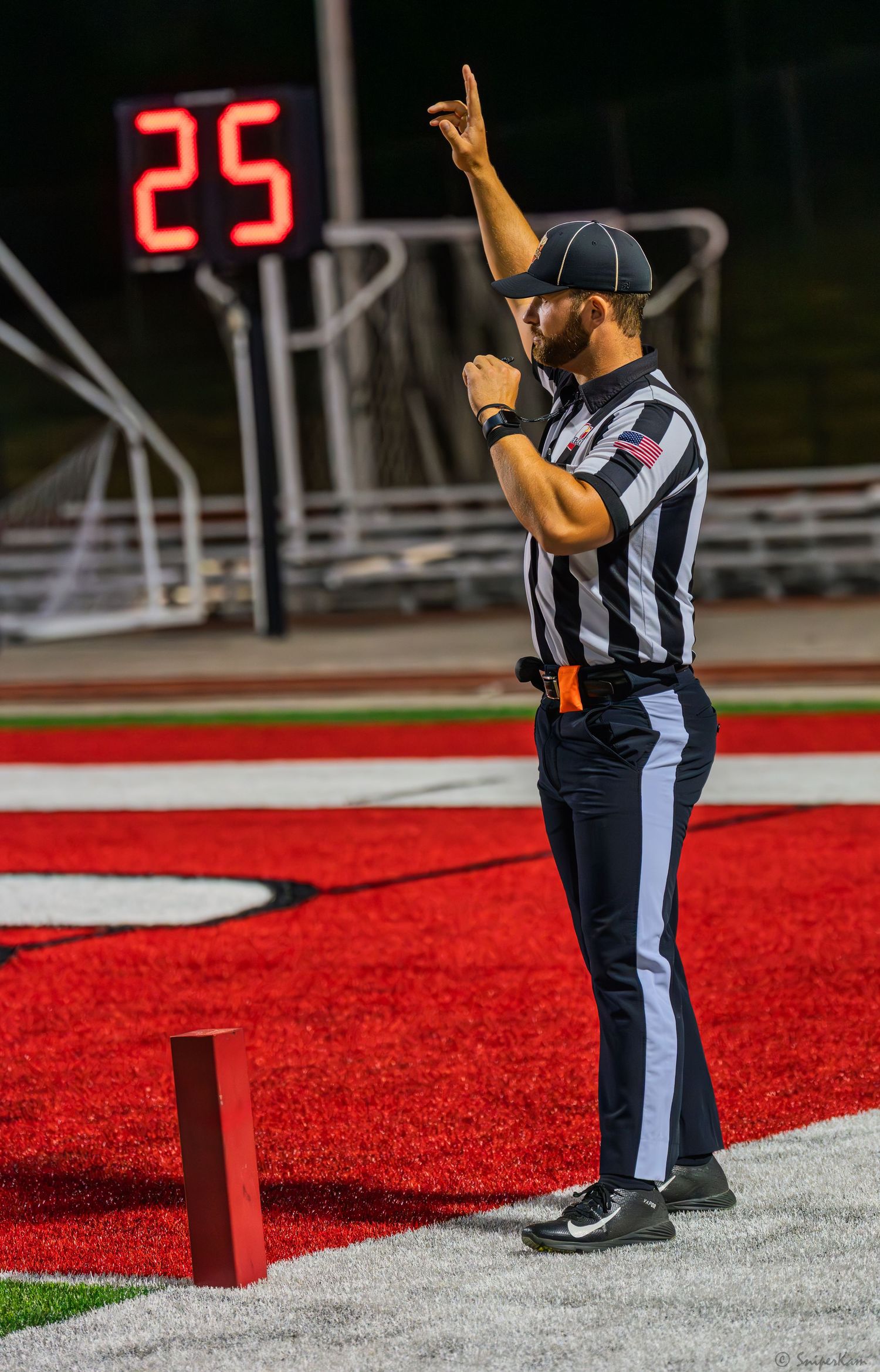 Football referee signals a call on red turf field at night with play clock showing 25 seconds.