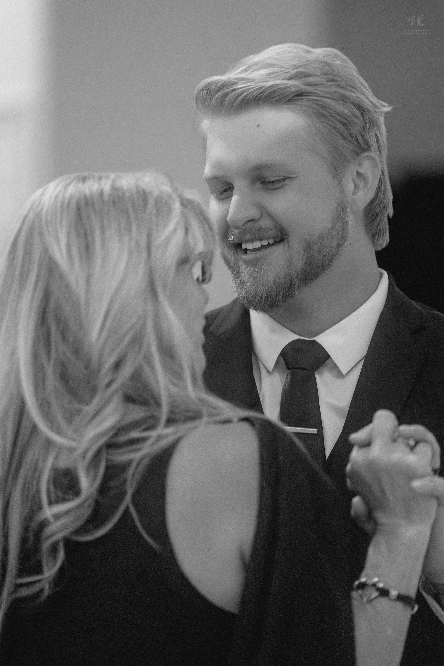 Smiling blond bearded groom in black suit shares intimate dance with woman at wedding reception.