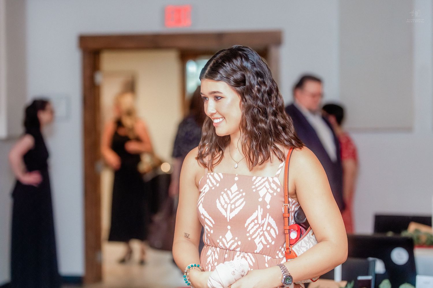 Smiling young woman in a floral pink dress at an indoor event venue with guests in the background.