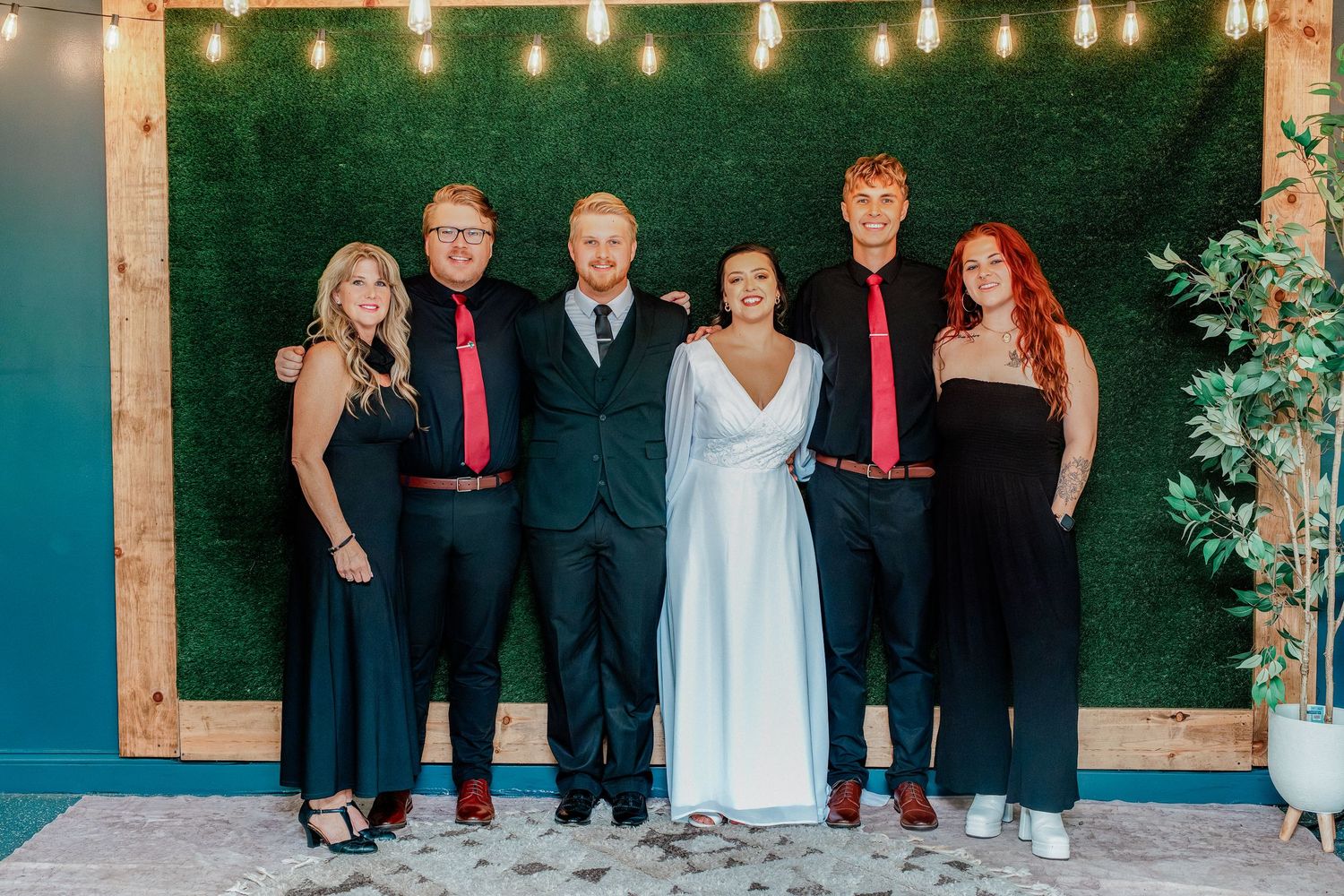 Six people posing together at a wedding, with bride and groom in center, groomsmen in black and red ties flanking them.