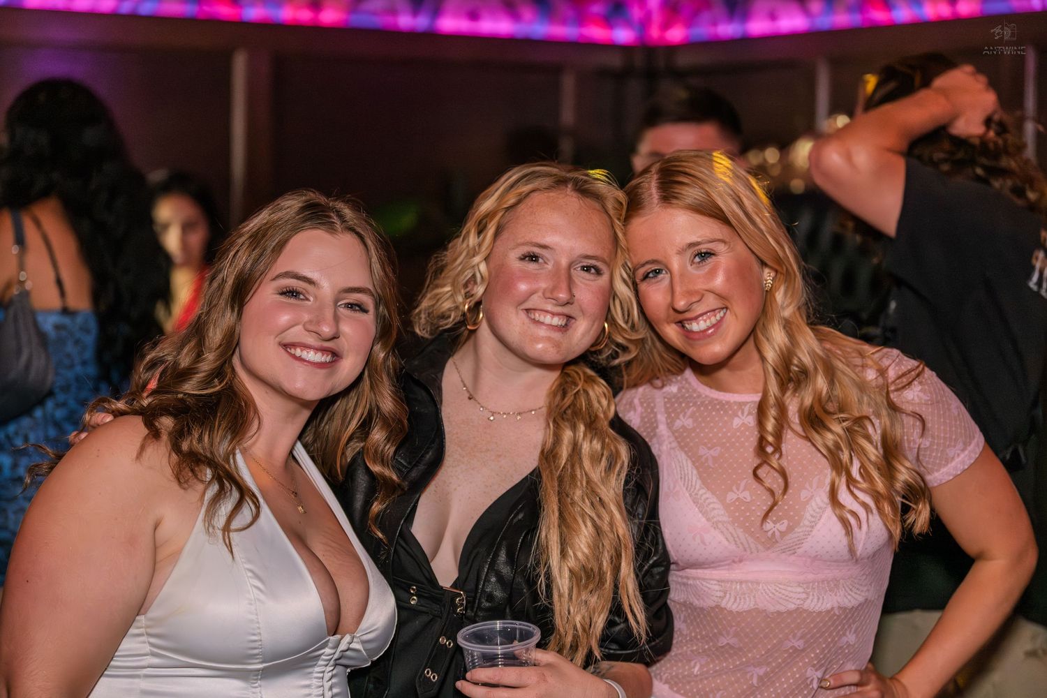Three smiling young women posing together at a nightclub under purple lights.