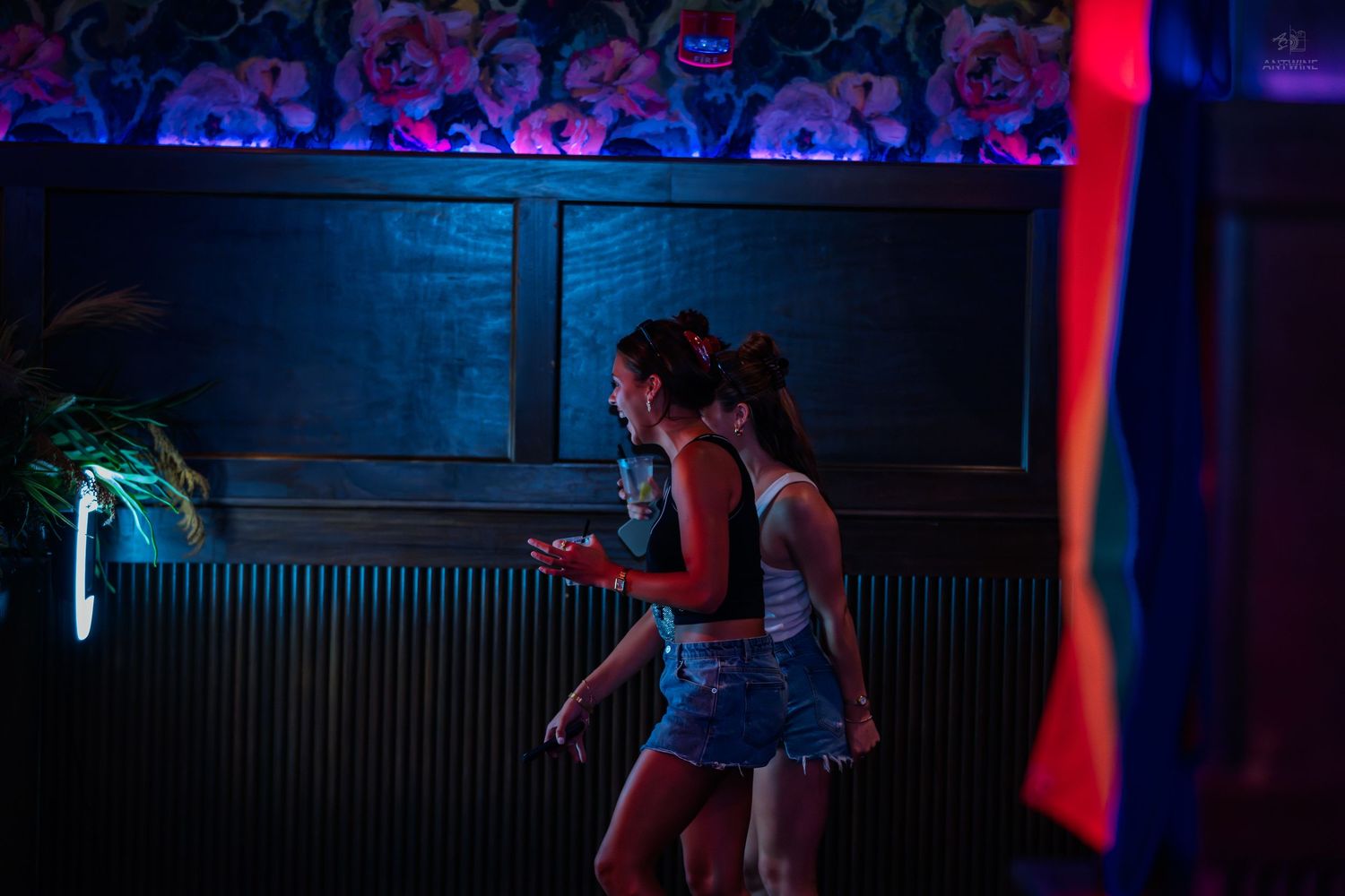 Two women walk through a dimly lit bar illuminated by blue and red neon lights.