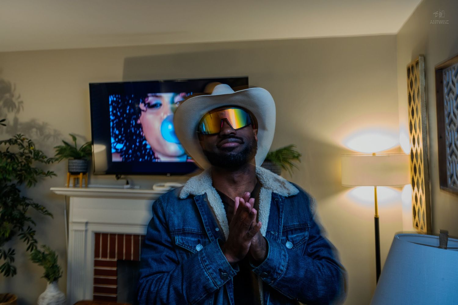 Man wearing white cowboy hat, mirrored sunglasses, and denim jacket with hands pressed together indoors.