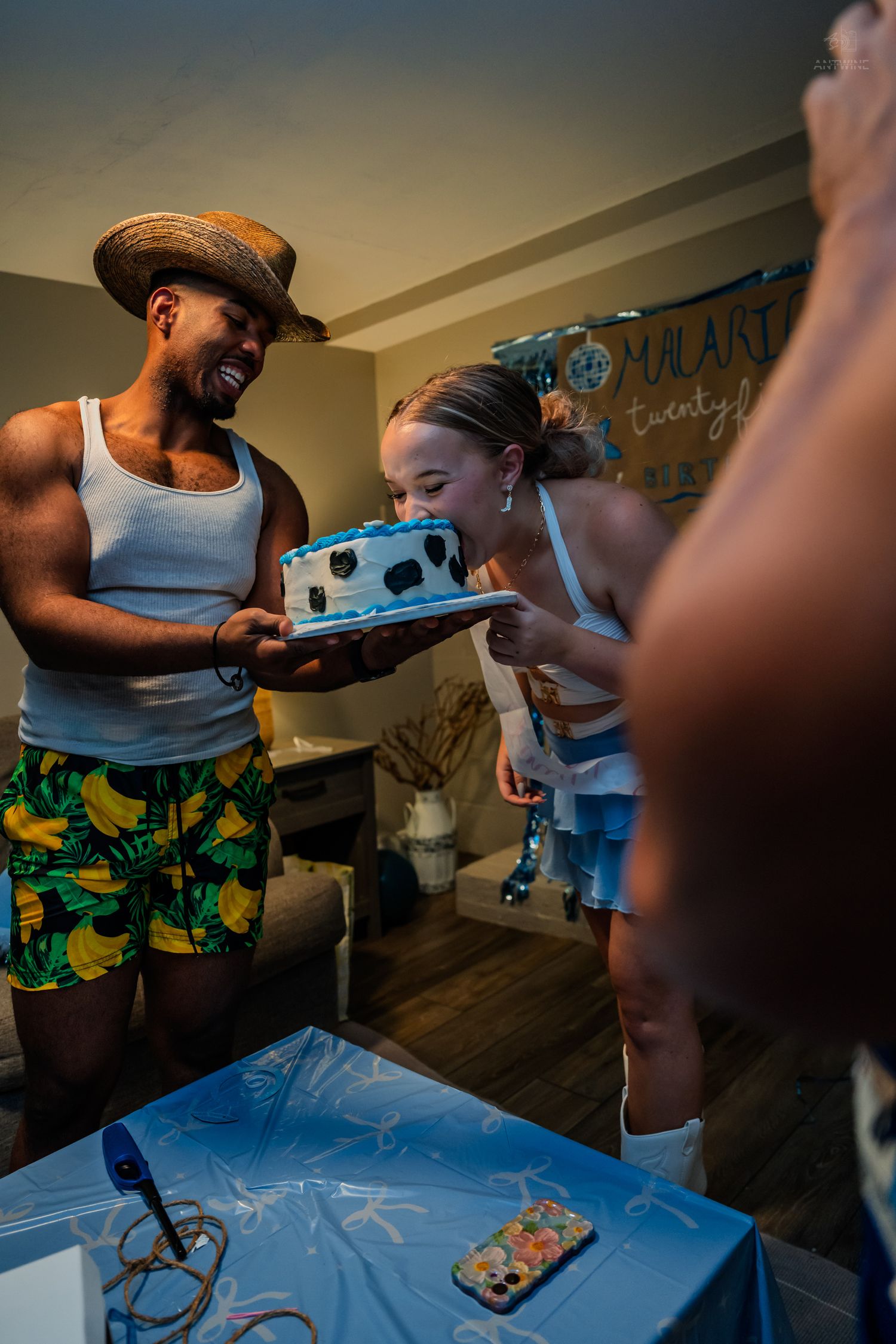 Man in straw hat holds blue birthday cake as woman leans in to take a bite at a party.