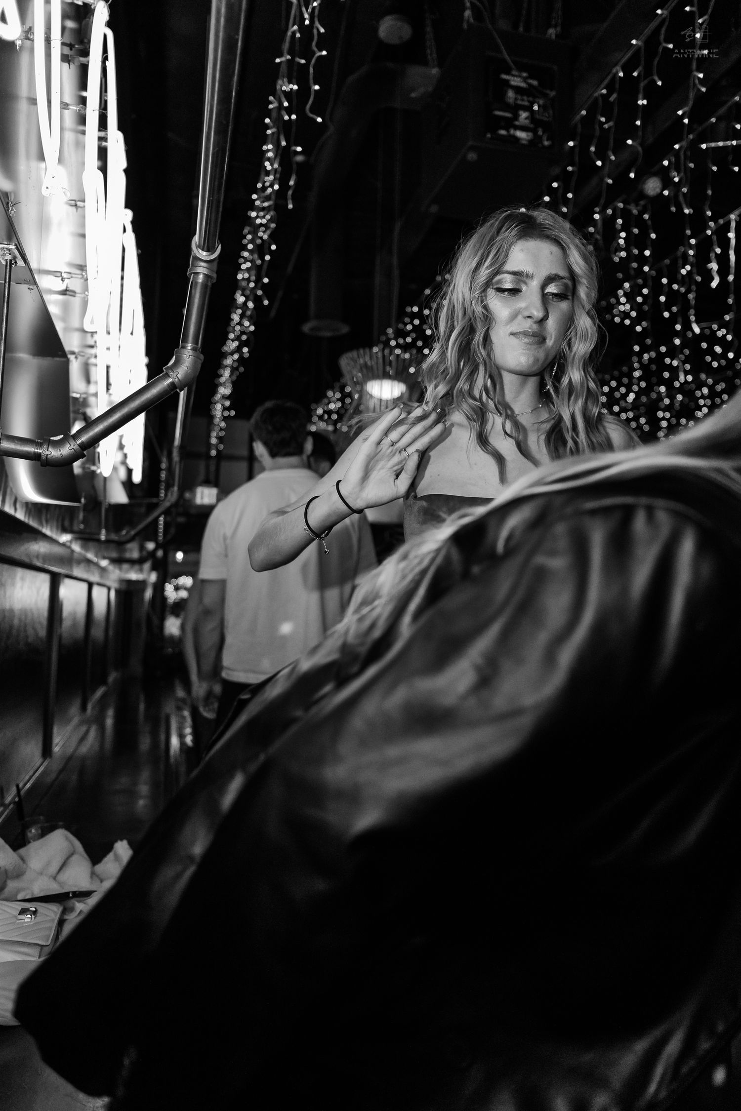 Young woman with wavy blonde hair holding a leather jacket in a venue decorated with string lights.