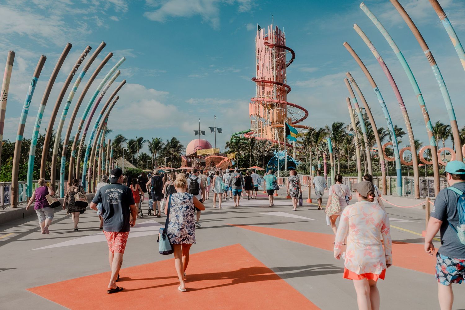 Tourists walk along a colorful promenade at a tropical theme park with a tall spiral ride tower.