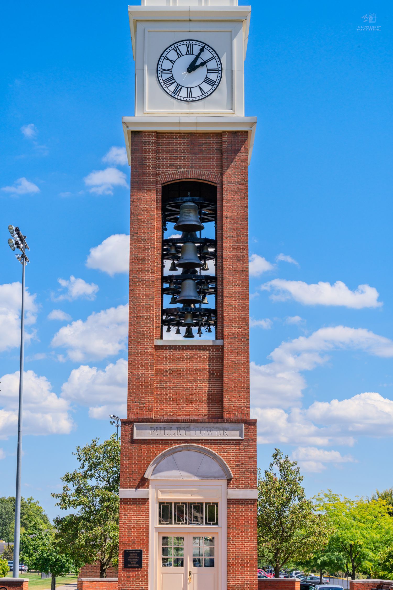 Tall red brick carillon bell tower with white clock and multiple bronze bells under blue sky.