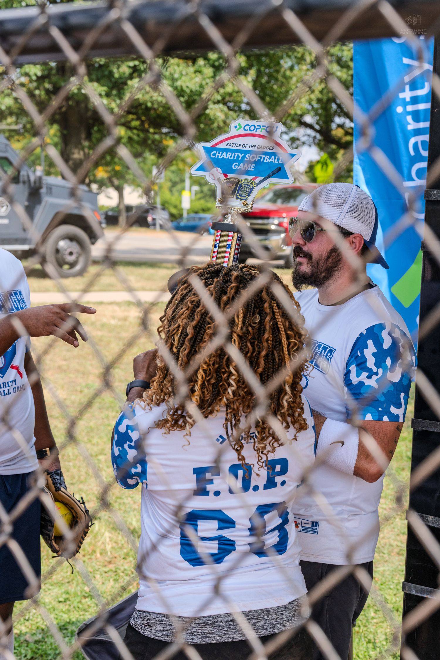 Players at a charity softball game examine a trophy behind a chain-link fence.