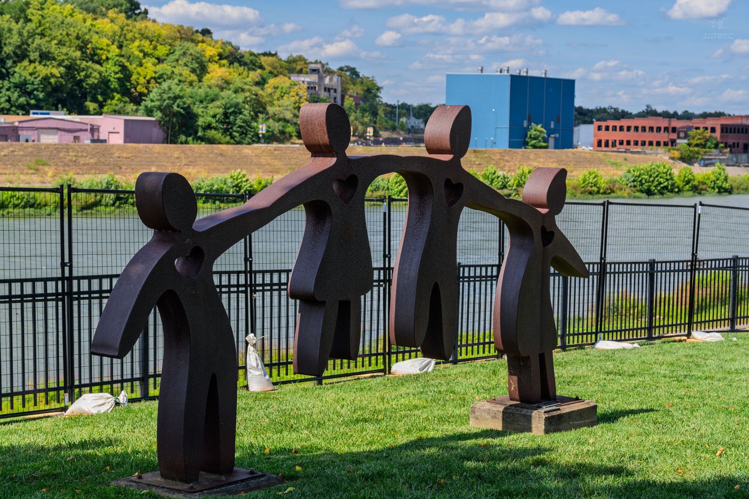 Abstract metal sculpture of three figures holding hands near a river with green landscape and buildings in background.