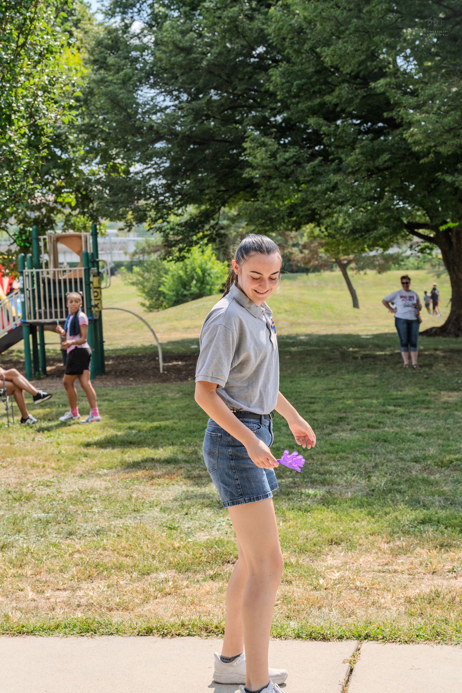 Smiling young woman in gray shirt and denim shorts holds a purple item in a sunny park setting.