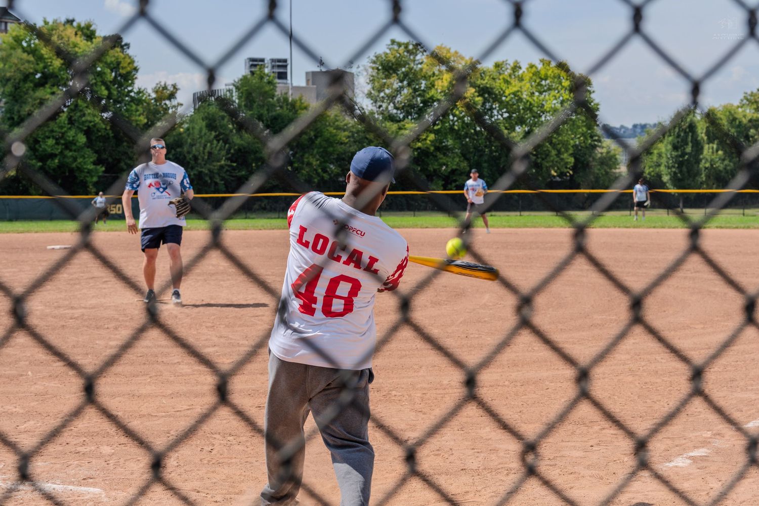 Softball batter in Local 48 jersey swinging at pitch viewed through chain-link fence.