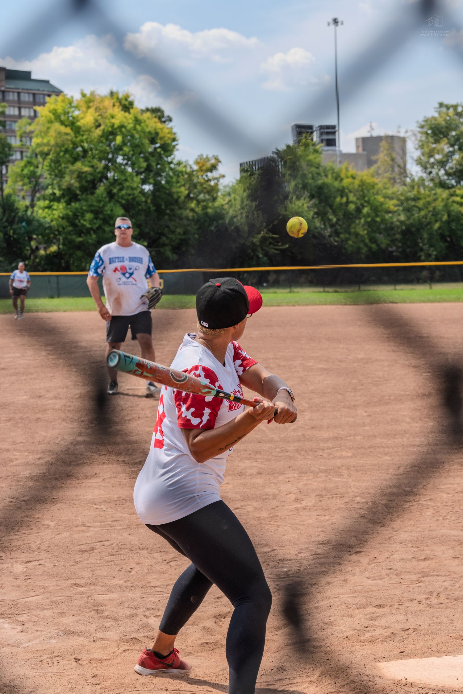 Batter in red jersey swings at yellow softball during outdoor game.