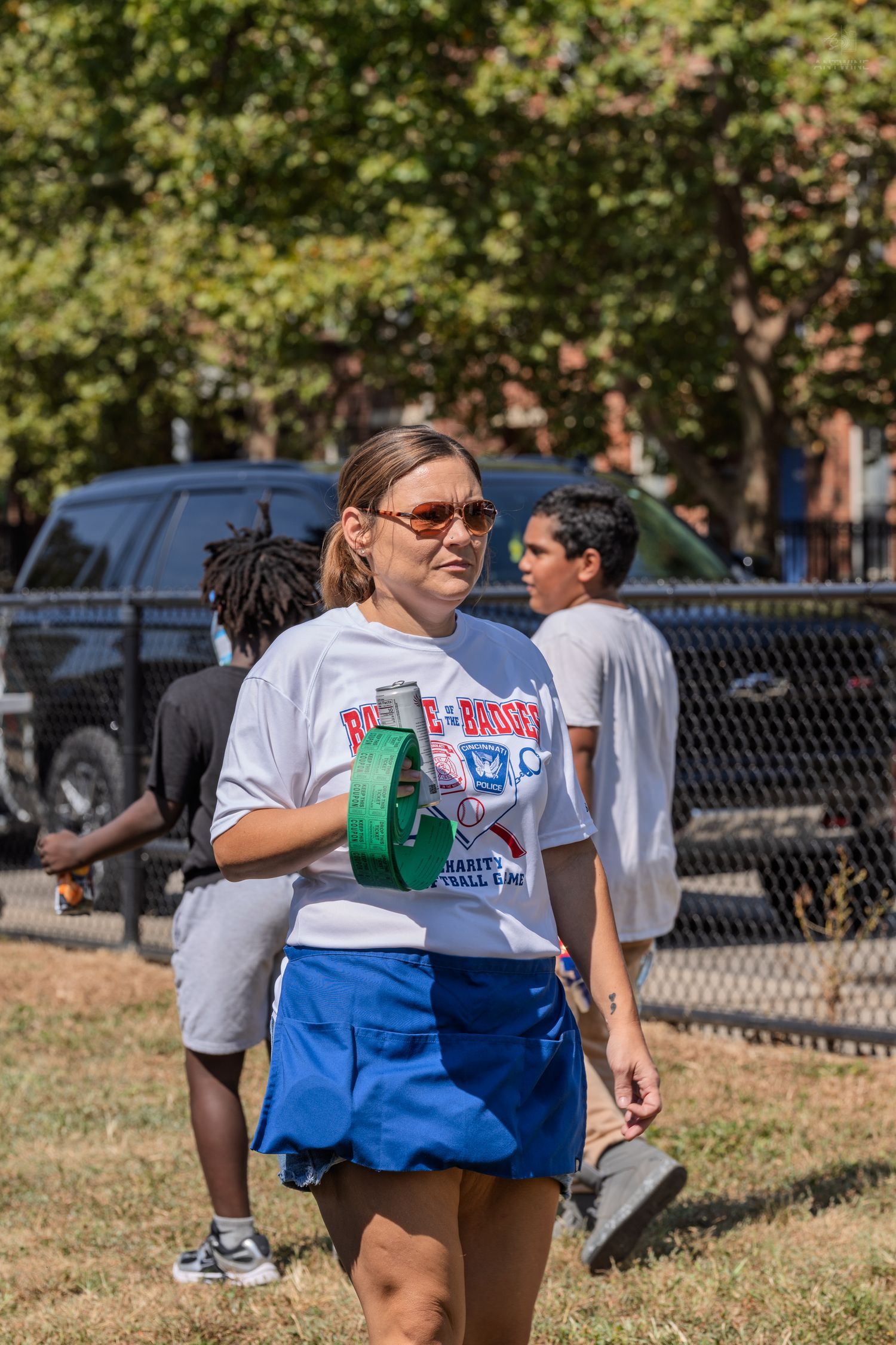 Woman in white graphic tee and blue skirt wearing sunglasses walks outdoors at a sunny park event.