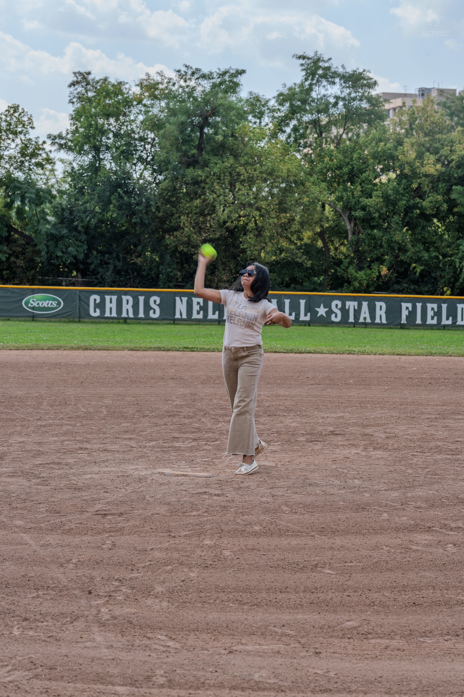 A woman in beige wide-leg pants throws a softball on Chris Nell All-Star Field during a ceremonial first pitch.