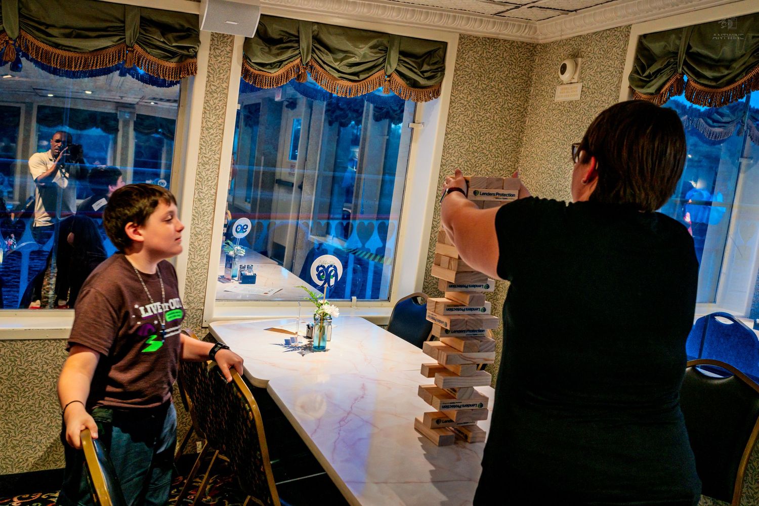 Two people play a Jenga tower game at a table in a softly lit banquet room.