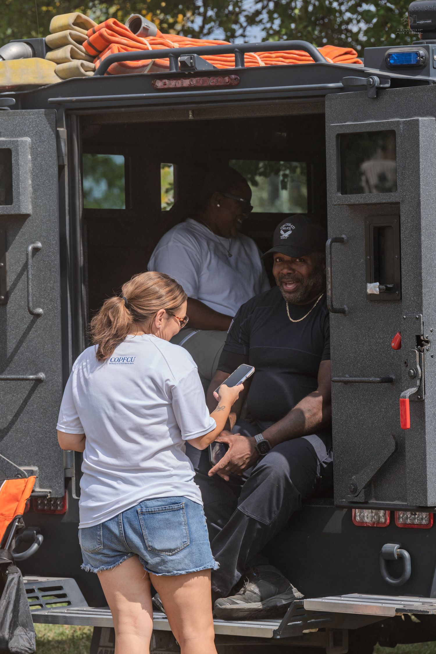 Two people chat with a smiling man seated in the open door of a tactical vehicle outdoors.